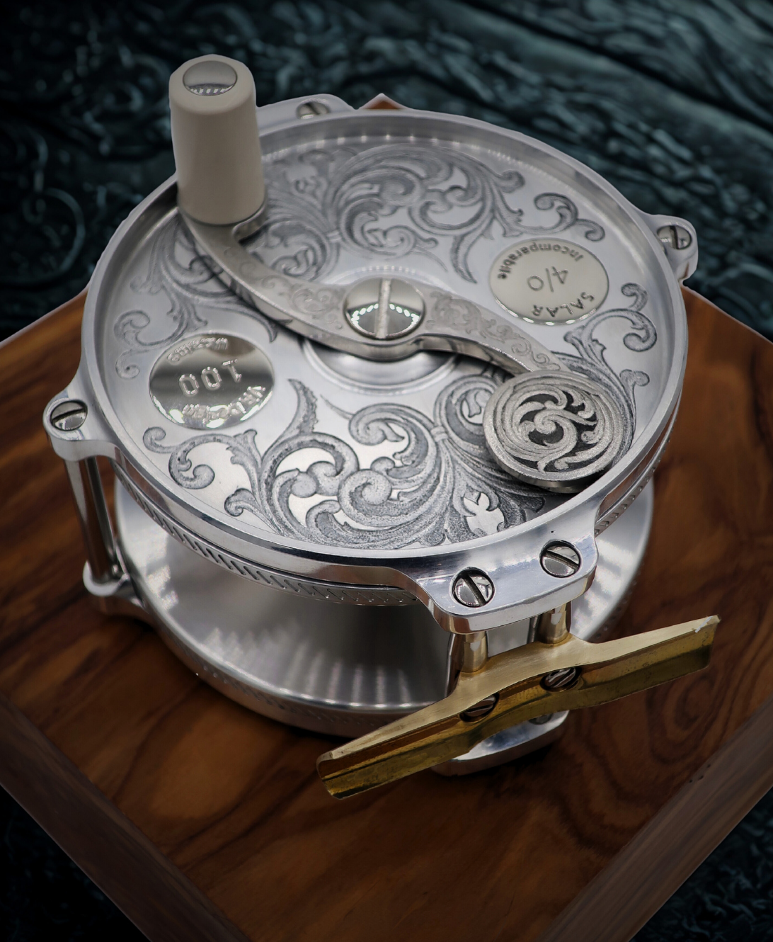 Hand engraved salar fly fishing reel by VRDesigns. The engraving features scrollwork. The reel is sitting on a piece  of wood.