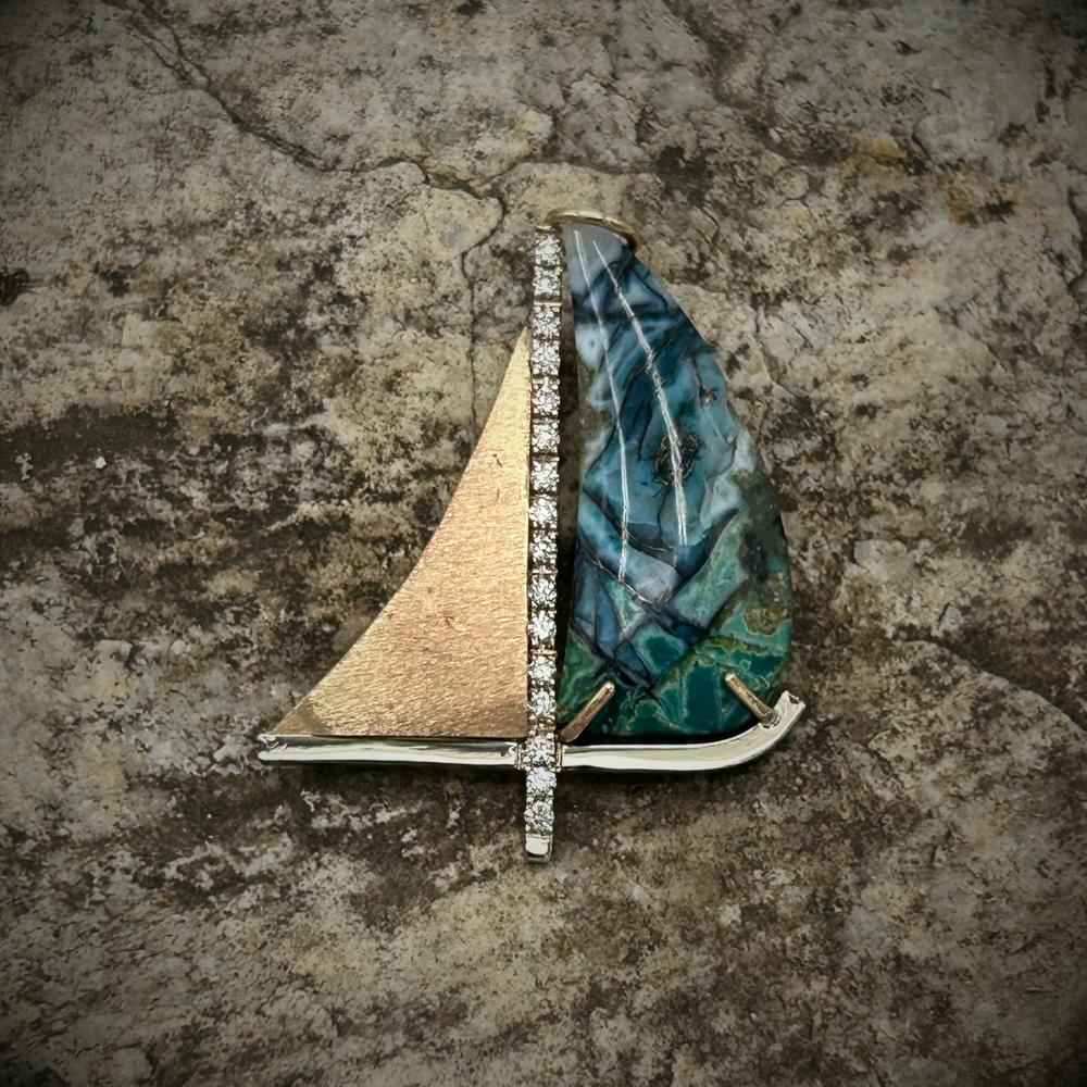Sailboat-shaped jewelry piece with gold sail and blue-green hull on a textured stone surface