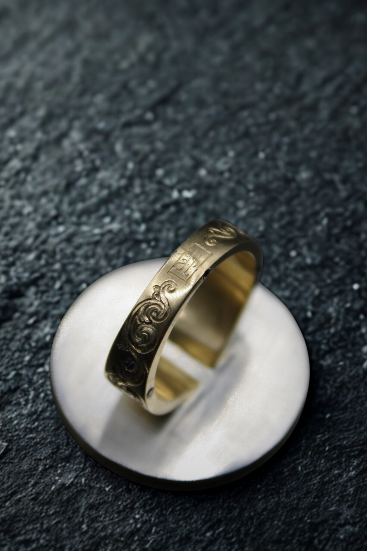 Gold ring with intricate design on a white stand against a dark textured background